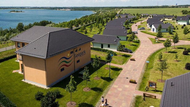 Et boligområde med en stor bygning, der hedder "Seepark Auerhain", med et tag bestemt af fliser, omgivet af velplejede græsplæner og en sti, der fører til et vandareal.