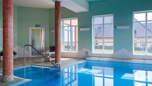 En swimmingpool med klart, blåt vand, omgivet af en flisbelagt terrasse. Store vinduer giver naturligt lys og udsigt udendørs.