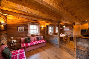 Ein gemütliches Blockhaus-Interieur mit Holzbalkendecke, einem Karo-Sofa, einem Esstisch und einem Kamin.