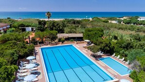A large outdoor swimming pool with multiple lanes, surrounded by lounge chairs and umbrellas, with a beach and ocean view in the background. - Residence Baia Verde, Valledoria (Valledoria)