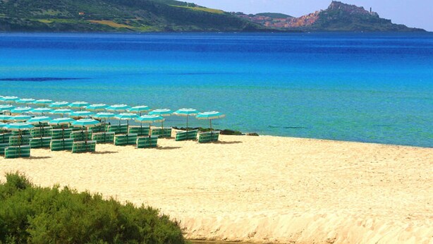 A beach with rows of green beach chairs and umbrellas, a sandy shore, and a distant hill with a structure on top. - Residence Baia Verde, Valledoria (Valledoria)