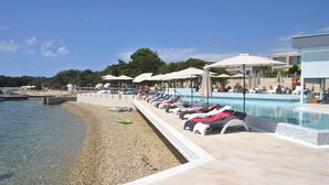 A poolside area with lounge chairs and umbrellas, a pebble beach, and a clear view of the sea. - Apartments in Villas Punta Skala With Beachpool (Zadar)
