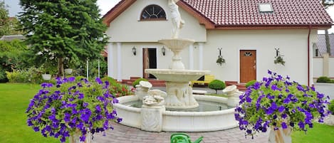 Uma casa com uma fonte, dois vasos de flores e um regador verde.