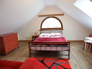 A bedroom with a sloped ceiling, a bed with a wooden headboard, a nightstand with a lamp, a dresser, and a window with a view. - Holiday Home Sobieszewo Near Baltic Sea (Gdańsk)