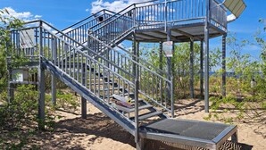 Una escalera metálica conduce a una plataforma techada, rodeada de arena y vegetación escasa.