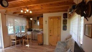 A cozy room with wooden floors, a dining table with chairs, a sofa, a TV, and a kitchen area with wooden cabinets and a clock on the wall. - Holiday Home Near Lake Mausz With Sauna (Parchowo)
