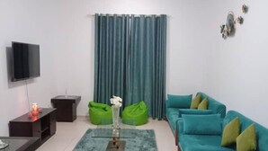 Een moderne woonkamer met een blauwgroene bank, groene relaxfauteuils, een flatscreen televisie en een glazen salontafel.