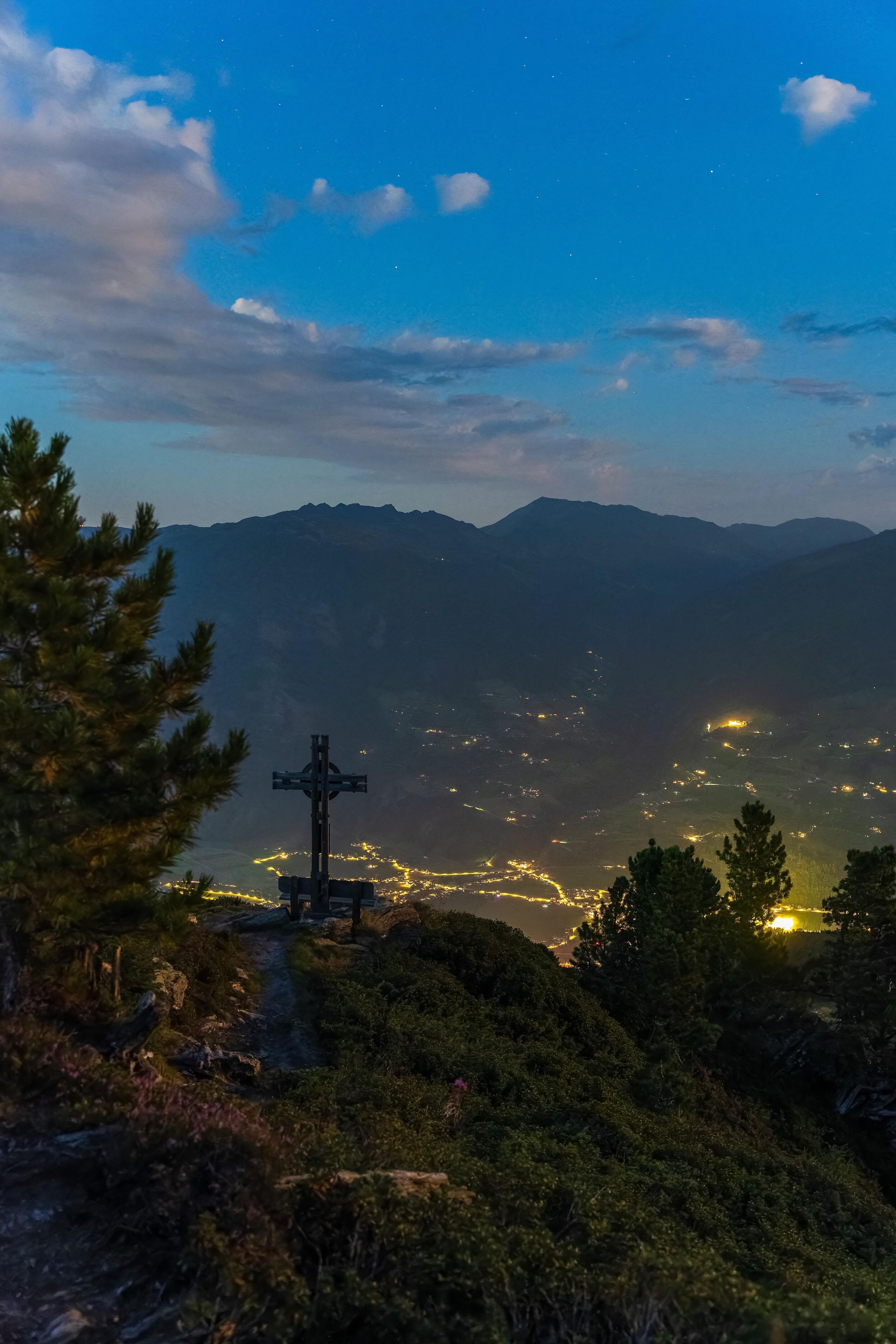 Ein Kruzifix auf einem Hügel mit Blick auf ein Tal und entfernte Stadtlichter.