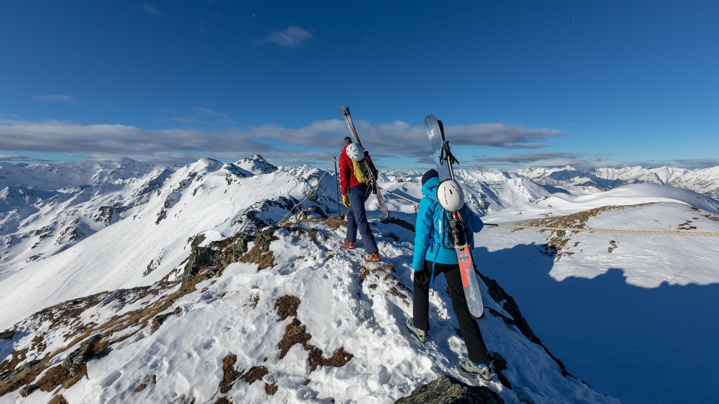 Paesaggio montano innevato con due sciatori in piedi su un crinale, che domina una vasta valle ricoperta di neve.