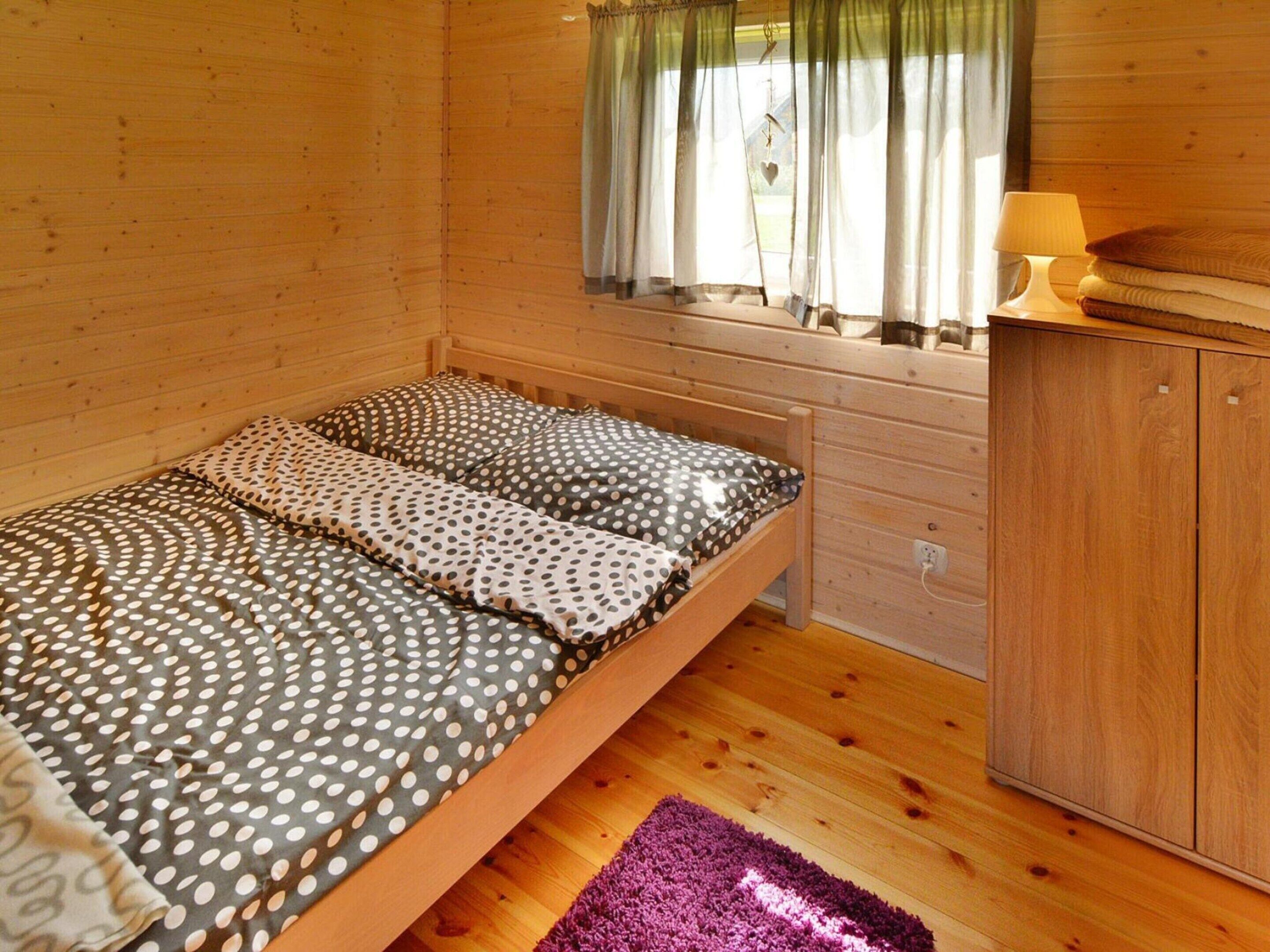 A wooden cabin room with a bed, a window with curtains, a nightstand with a lamp, and a wooden wardrobe.