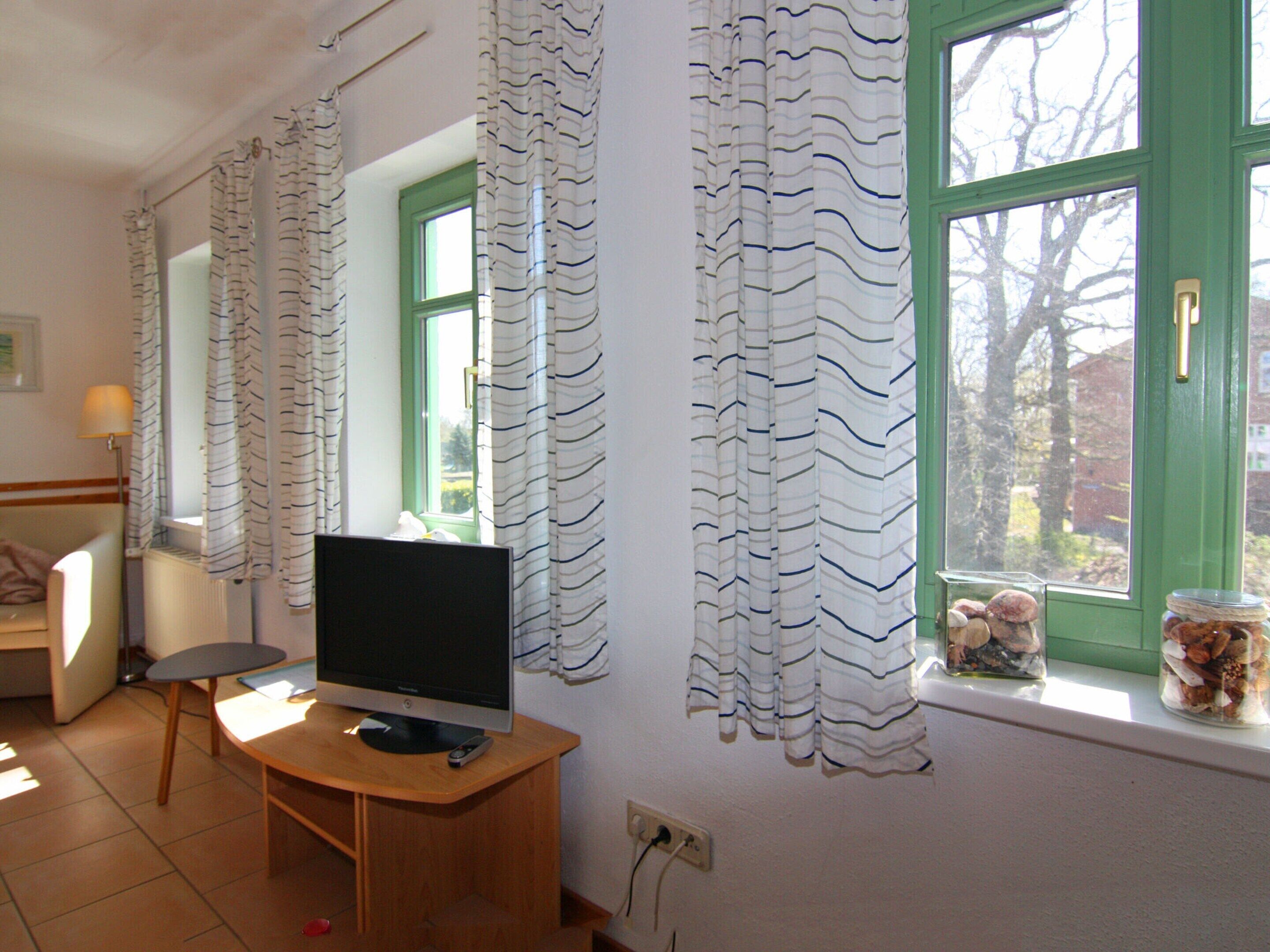 A room with a TV on a wooden desk, a chair, a window with green shutters, and a wall-mounted lamp.
