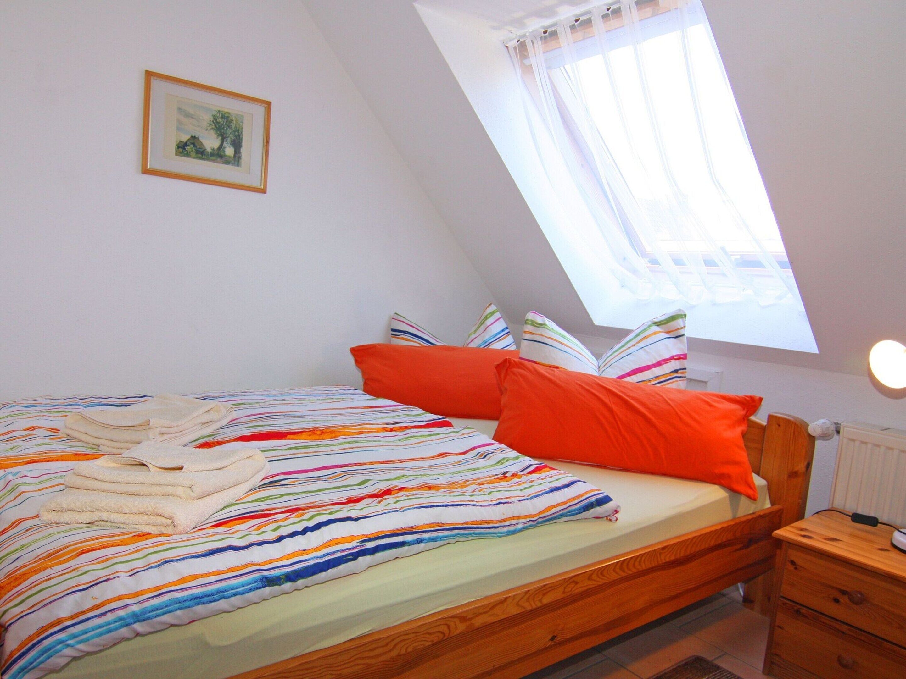 A bedroom with a bed, pillows, a nightstand, a wooden headboard, a window with sheer curtains, and a framed picture on the wall.