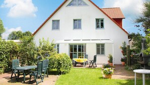 A two-story house with a red-tiled roof, a covered patio with a yellow table, and a garden with various plants and a barbecue grill. - Appartementhaus Kranichblick, Breege (Breege)