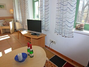 A room with a bed, a desk with a computer, a chair, a table with bottles, and a window with curtains. - Appartementhaus Kranichblick, Breege (Breege)