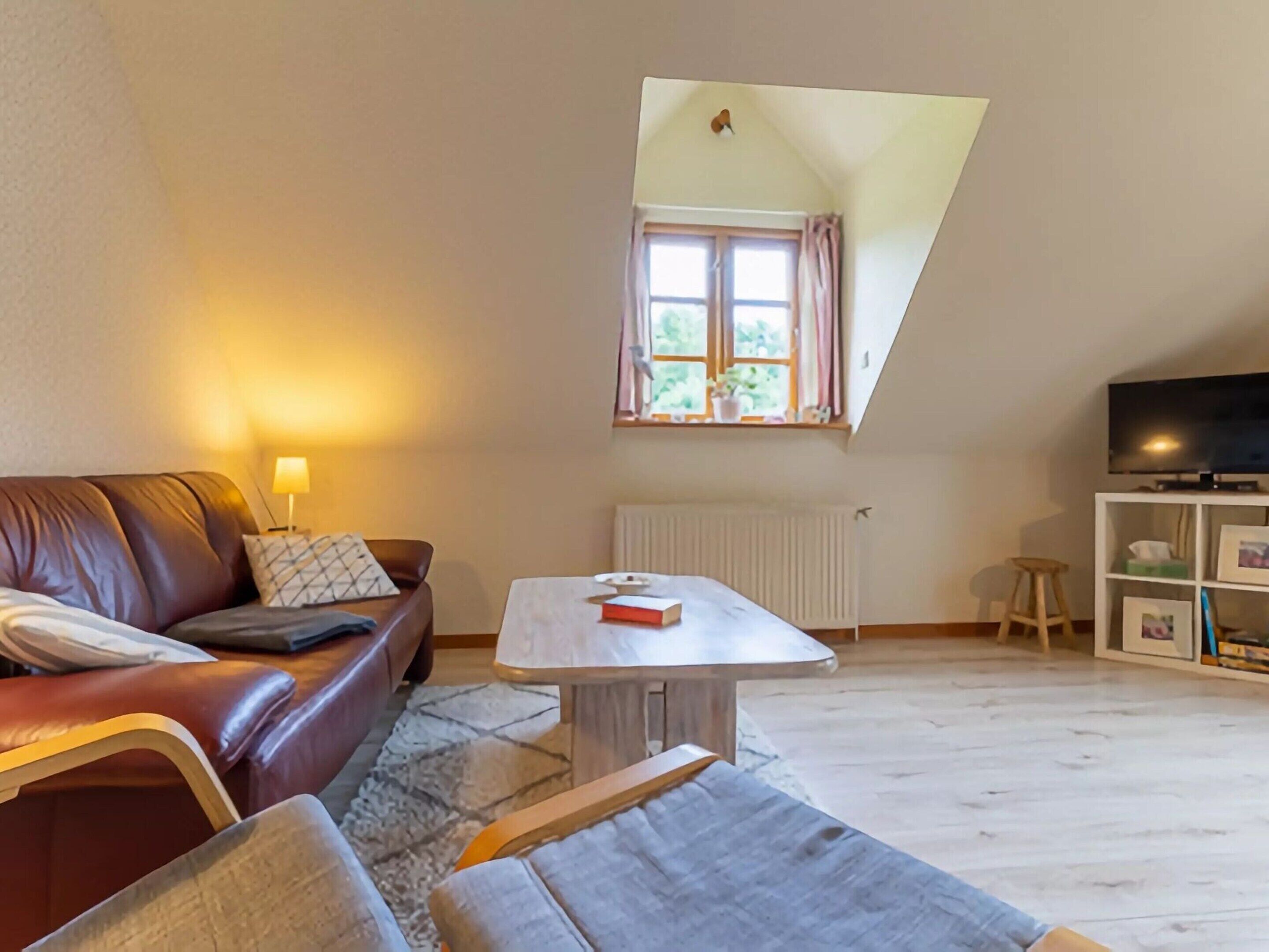 Ein Wohnzimmer mit braunem Ledersofa, einem kleinen Tisch, einem Fernseher auf einem Schrank und einem Fenster mit Vorhängen.