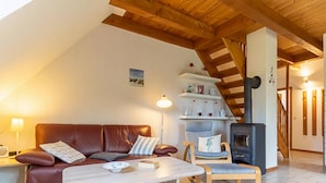 A cozy living room with a brown leather sofa, a wooden coffee table, and a blue armchair. A wooden staircase leads to a loft area with a fireplace. - Landhaus in Vollerwiek (Vollerwiek)