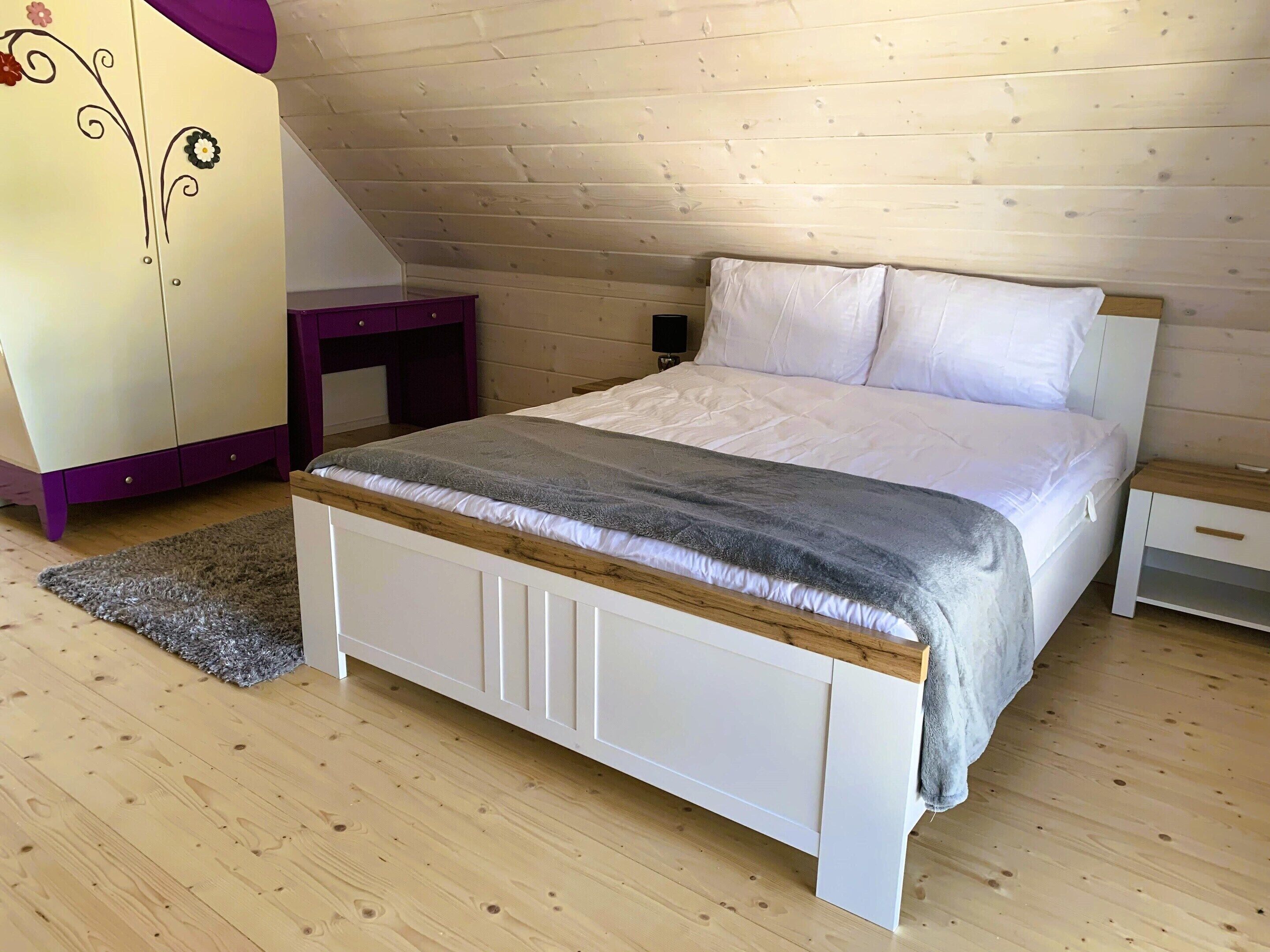 A bedroom with a wooden sloped ceiling, a bed with white bedding, a purple dresser, and a small wooden nightstand.
