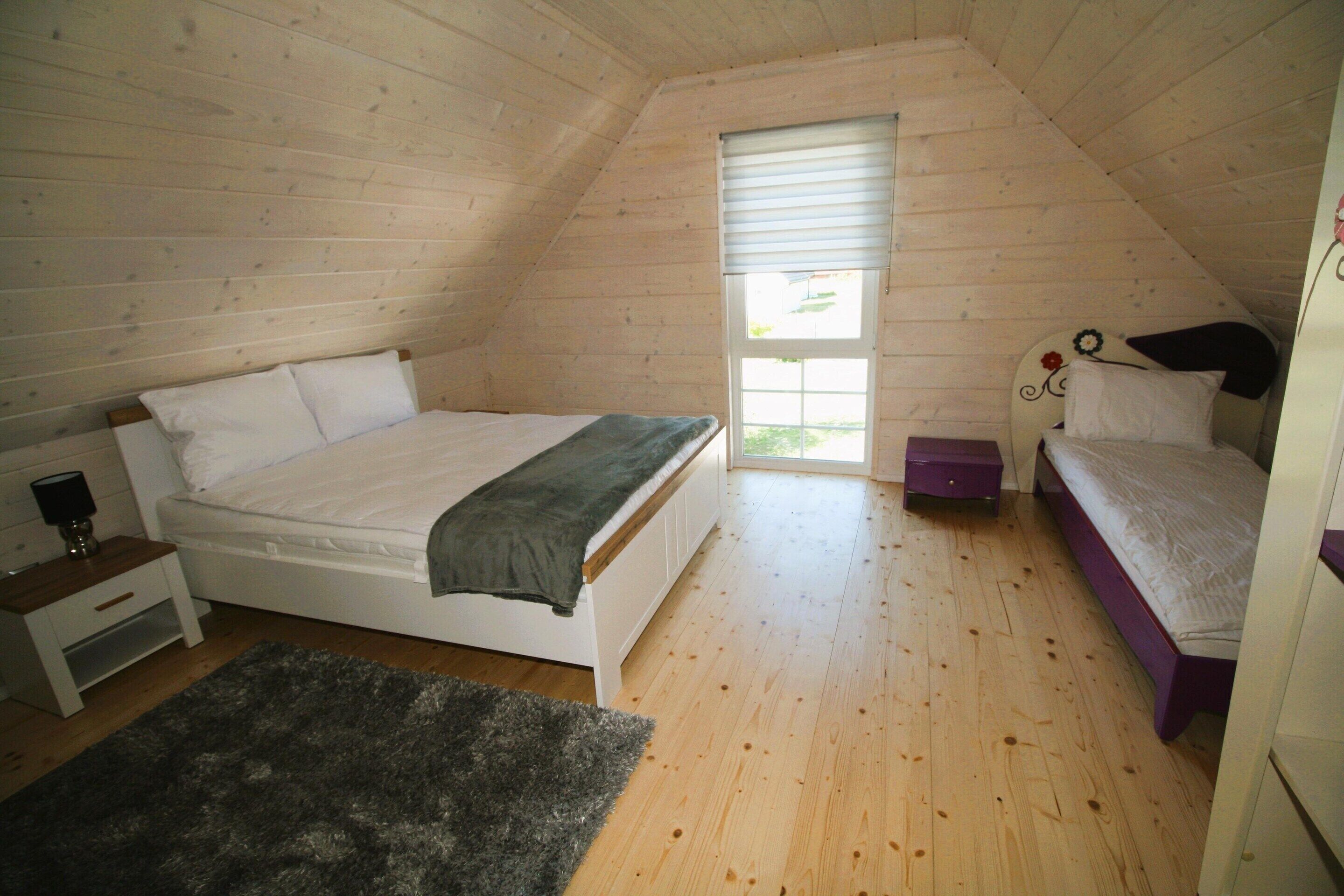 A bedroom with a sloped ceiling, a bed with a white mattress and a dark blanket, a small bedside table with a lamp, and a window with blinds.