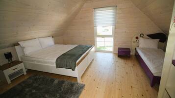 A bedroom with a sloped ceiling, a bed with a white mattress and a dark blanket, a small bedside table with a lamp, and a window with blinds.