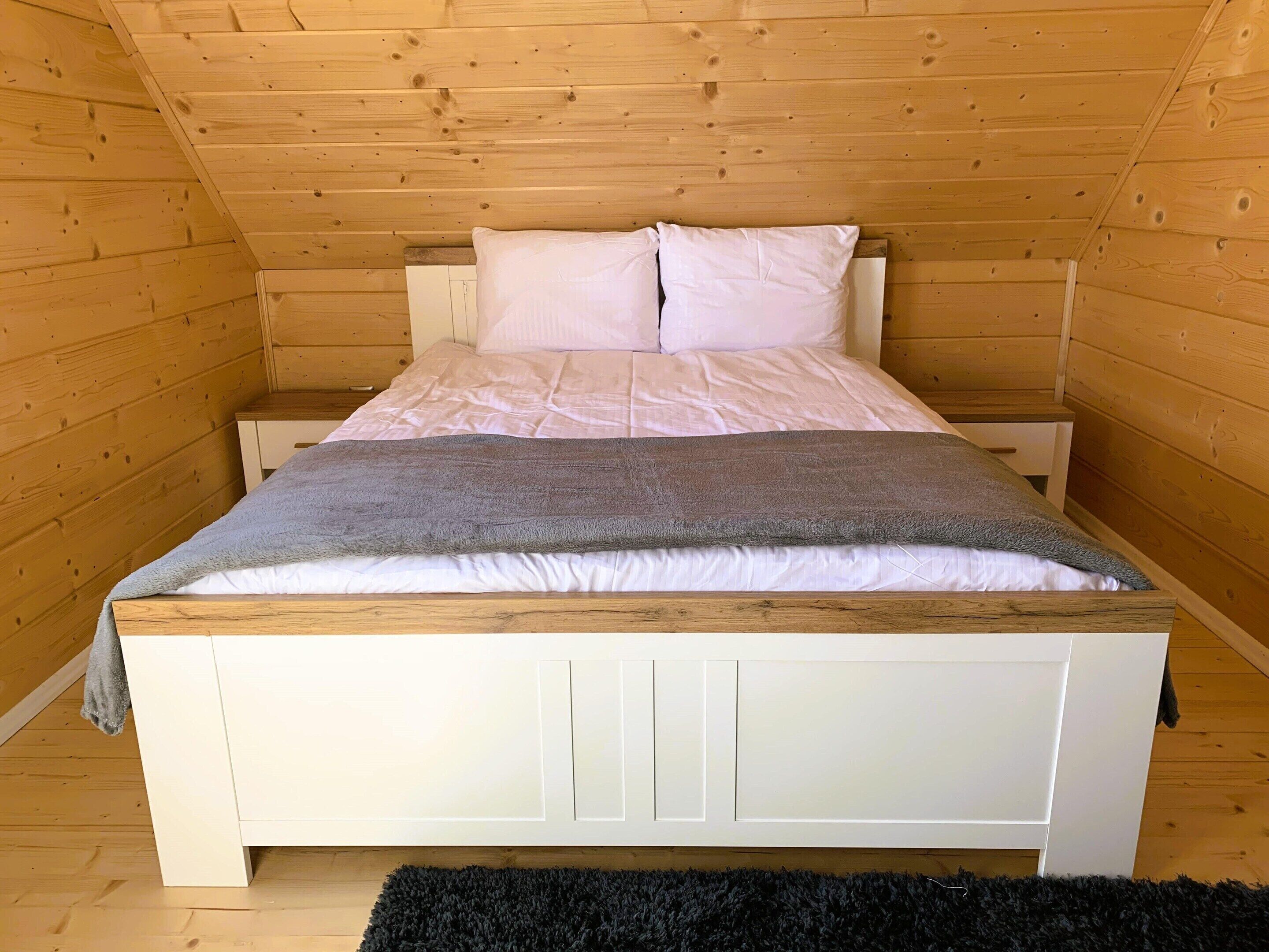A wooden-paneled room with a bed, white nightstands, and a black rug.