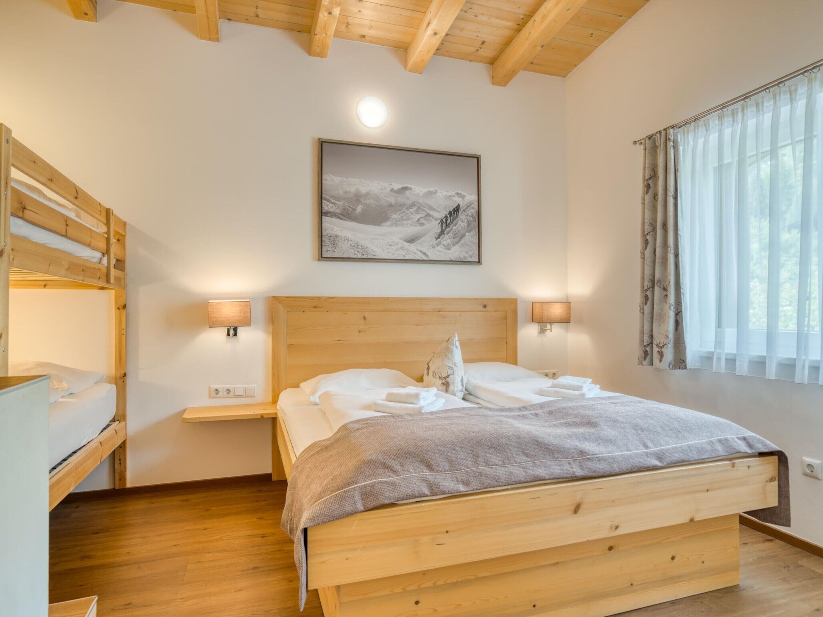Habitación con literas de madera, una cama individual, dos lámparas de noche, un cuadro en la pared y una ventana con cortinas.