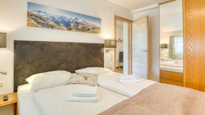 A hotel room with a large bed, a headboard, a painting of mountains, a wooden wardrobe, and a mirror. - Chalet in Neukirchen With Mountain Views (Neukirchen am Grossvenediger)