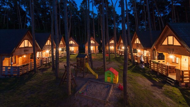 Eine Reihe aus Holzgebäuden mit einem Spielplatz in der Mitte, umgeben von hohen Bäumen.