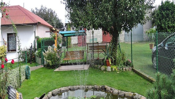 Un estanque con nenúfares y una estatua de pájaro en un jardín con valla, banco y un edificio al fondo.