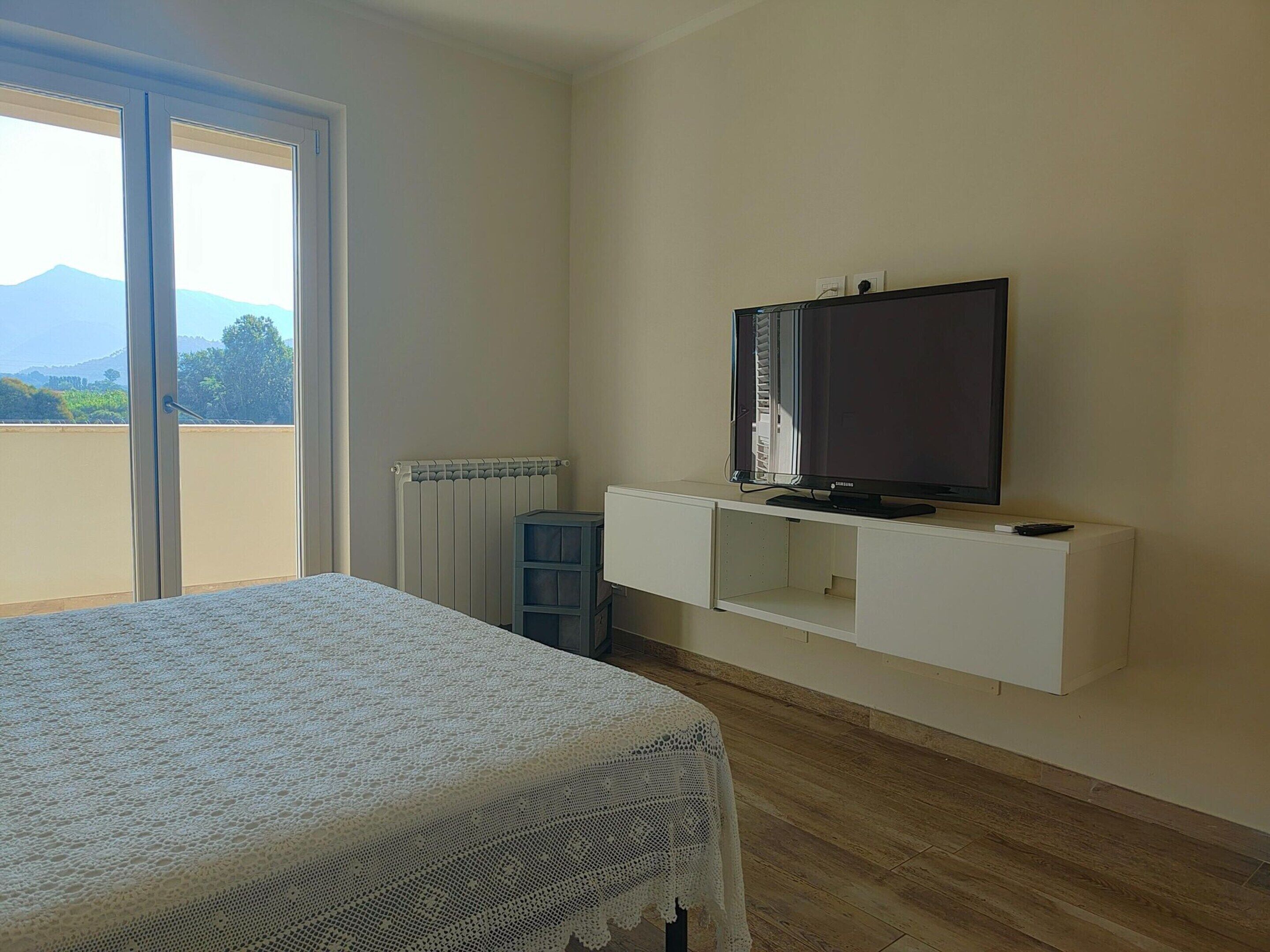Un dormitorio con cama, televisor montado en la pared, radiador y vistas al exterior.