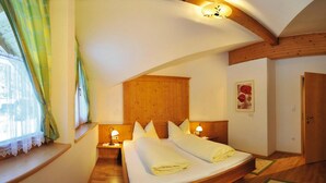 Un dormitorio con cabecera de madera, dos mesitas de noche con lámparas, una cama grande con mantas ordenadas, una ventana con cortinas y una puerta de madera.