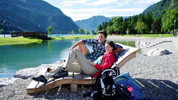 Una pareja descansando en un banco de madera junto a un lago, con montañas al fondo.