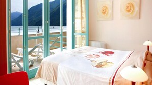 A hotel room with a bed, a red chair, a lamp, and a view of mountains through large windows. - Modern Porlezza Apartment for 4 (Porlezza)