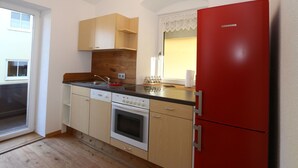 A compact kitchen with wooden cabinets, a red refrigerator, a built-in oven, and a window with lace curtains. - Apartment in Fugen Near Spieljochbahn (Fuegen)