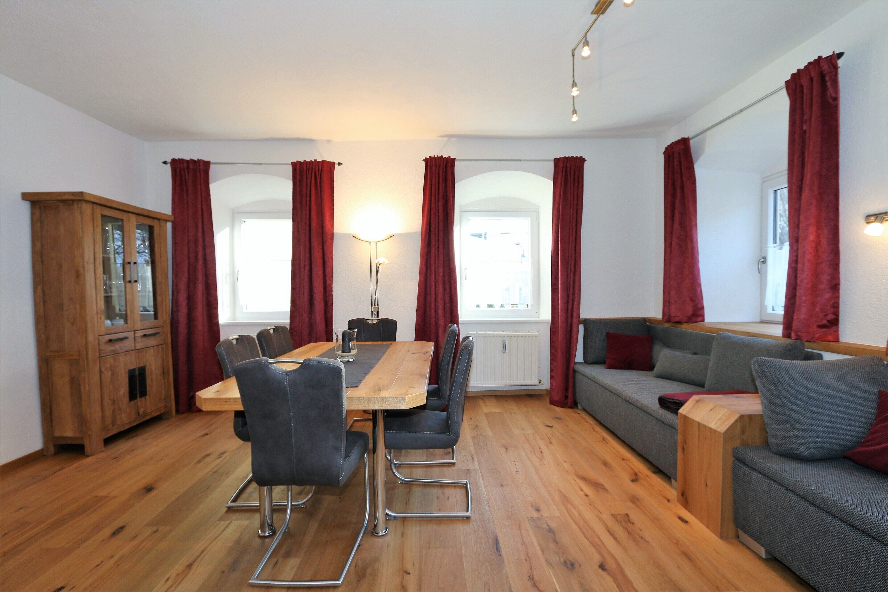 A modern living room with a wooden dining table, grey sofa, and red curtains.