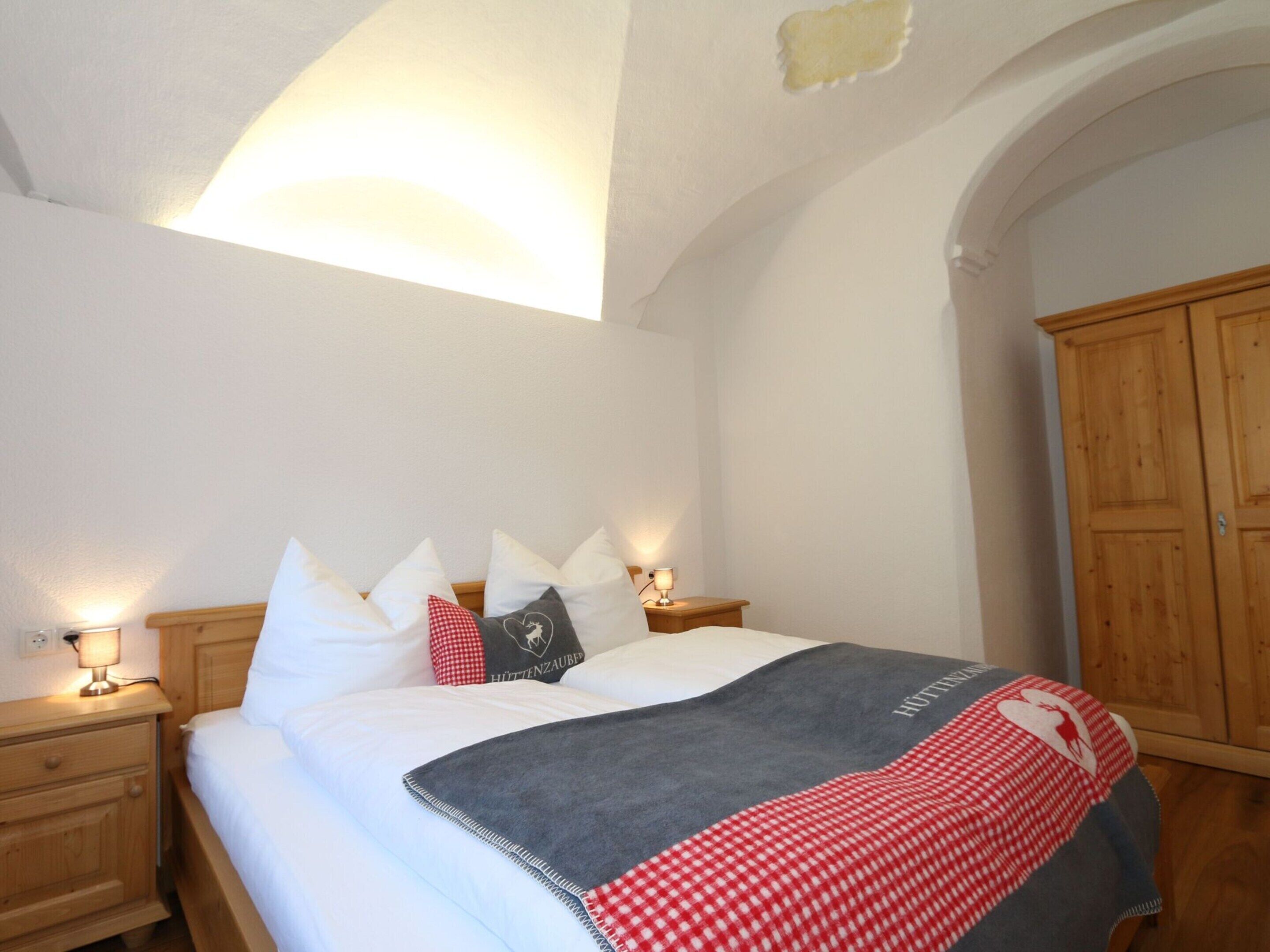 A bedroom with a bed, pillows, a blanket, a nightstand with a lamp, and a wooden wardrobe.