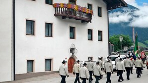 A building with a balcony adorned with colorful flowers, a group of people in uniform walking in front, and a mountainous backdrop. - Apartment in Fugen Near Spieljochbahn (Fuegen)