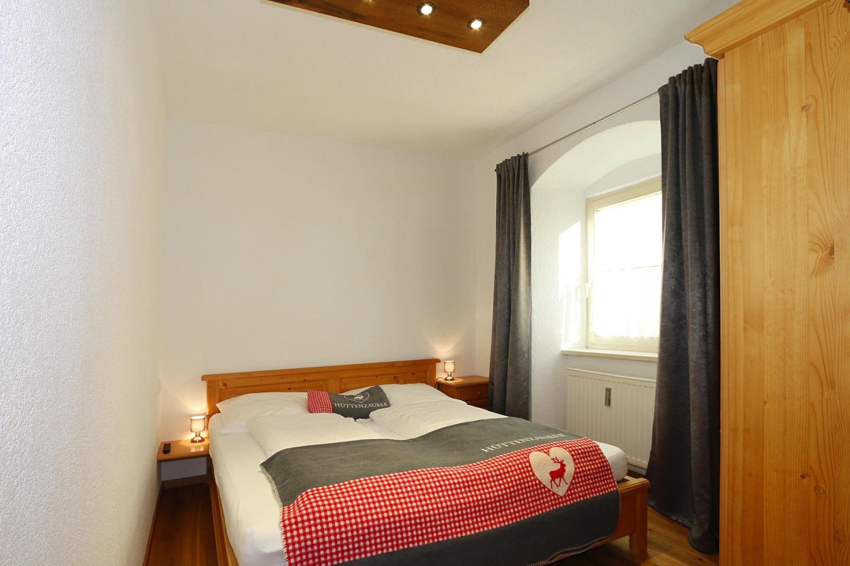 Une chambre d’hôtel avec un lit en bois, une table de chevet avec une lampe, une fenêtre avec des rideaux et une armoire en bois.