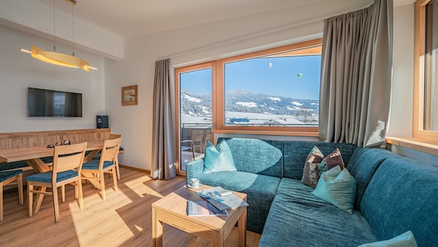 Un salon moderne avec une table à manger en bois, un canapé bleu et une grande fenêtre donnant sur un paysage montagneux enneigé.