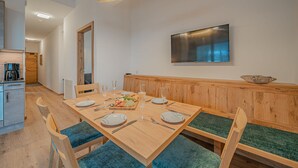 Un coin repas avec une table en bois dressée pour quatre personnes, des bancs rembourrés de couleur verte, un luminaire suspendu moderne et un téléviseur à écran plat fixé au mur.