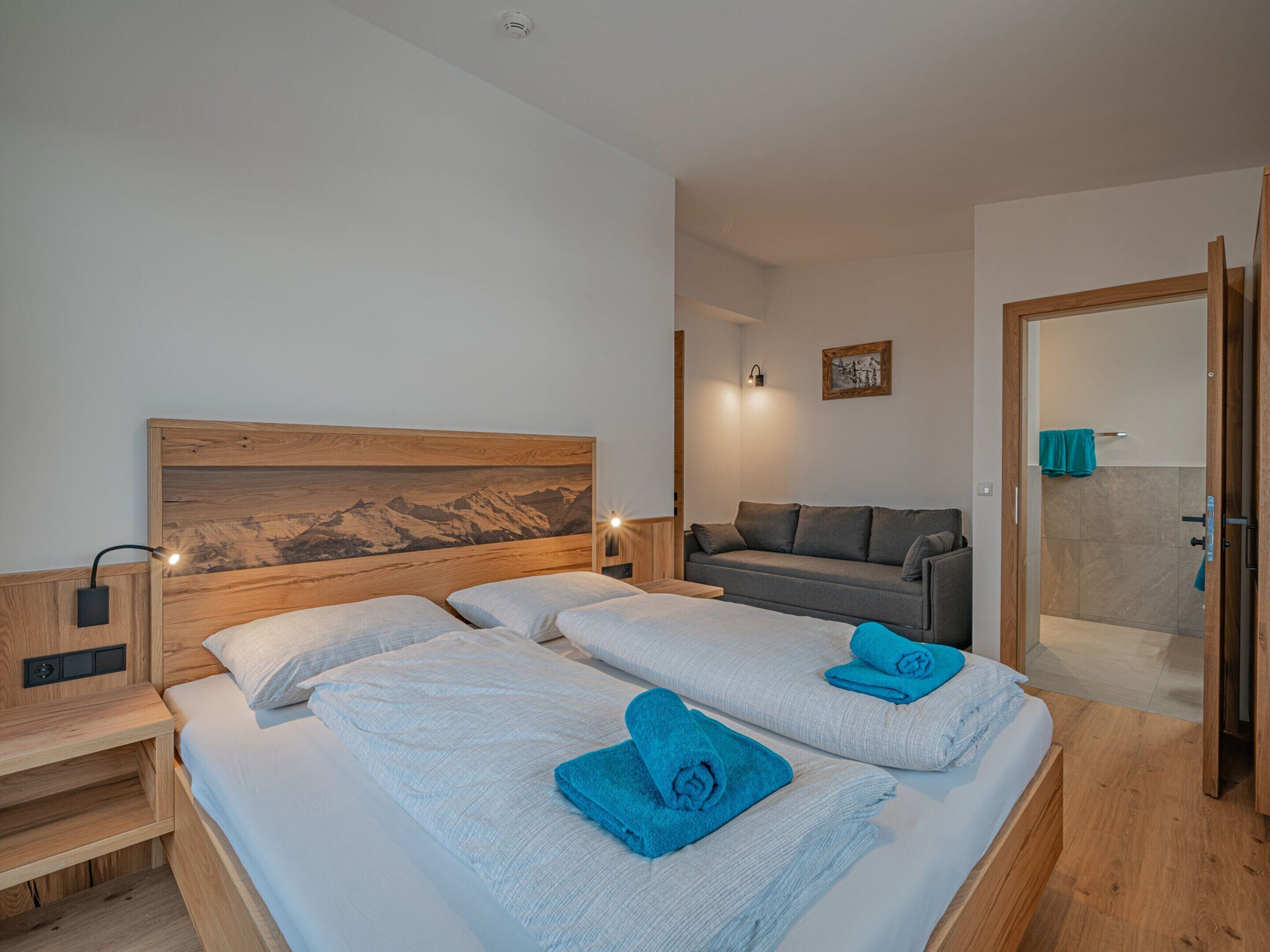 Habitación de hotel con dos camas, un sofá, un cabecero de madera, una lámpara fijada a la pared y una puerta que da a un baño.