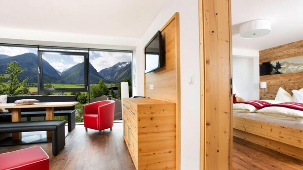 A modern room with a wooden desk, a red chair, a bed with a mountain view, and a TV mounted on the wall. - Apartment in Hohe Tauern With Mountain Views (Neukirchen am Grossvenediger)
