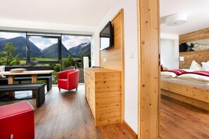Een moderne kamer met een houten bureau, een rode stoel, een bed met uitzicht op de bergen, en een aan de muur bevestigde televisie.