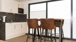 A modern kitchen with white cabinets, a dark countertop, and a large window. There are two brown bar stools and a wooden table. - Hausboot in Boltenhagen (Boltenhagen)