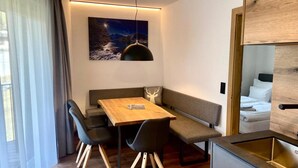 A modern hotel room with a wooden dining table, black chairs, a bench, a large window with sheer curtains, a painting of a night scene, and a kitchenette with a sink. - Bergzeit Apartments Near ski Slope (Saalbach-Hinterglemm)