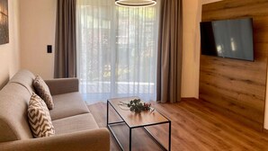 A modern living room with a sofa, a coffee table with decorative items, a TV mounted on a wooden wall, and large windows with sheer curtains. - Bergzeit Apartments Near ski Slope (Saalbach-Hinterglemm)