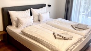 A double bed with white linens and two pillows, a headboard, a bedside table, and a painting of a mountain landscape on the wall. - Bergzeit Apartments Near ski Slope (Saalbach-Hinterglemm)