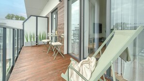 Un balcón moderno con cubierta de madera, una zona de comedor con sillas y una mesa, y una silla de lounge con una manta.