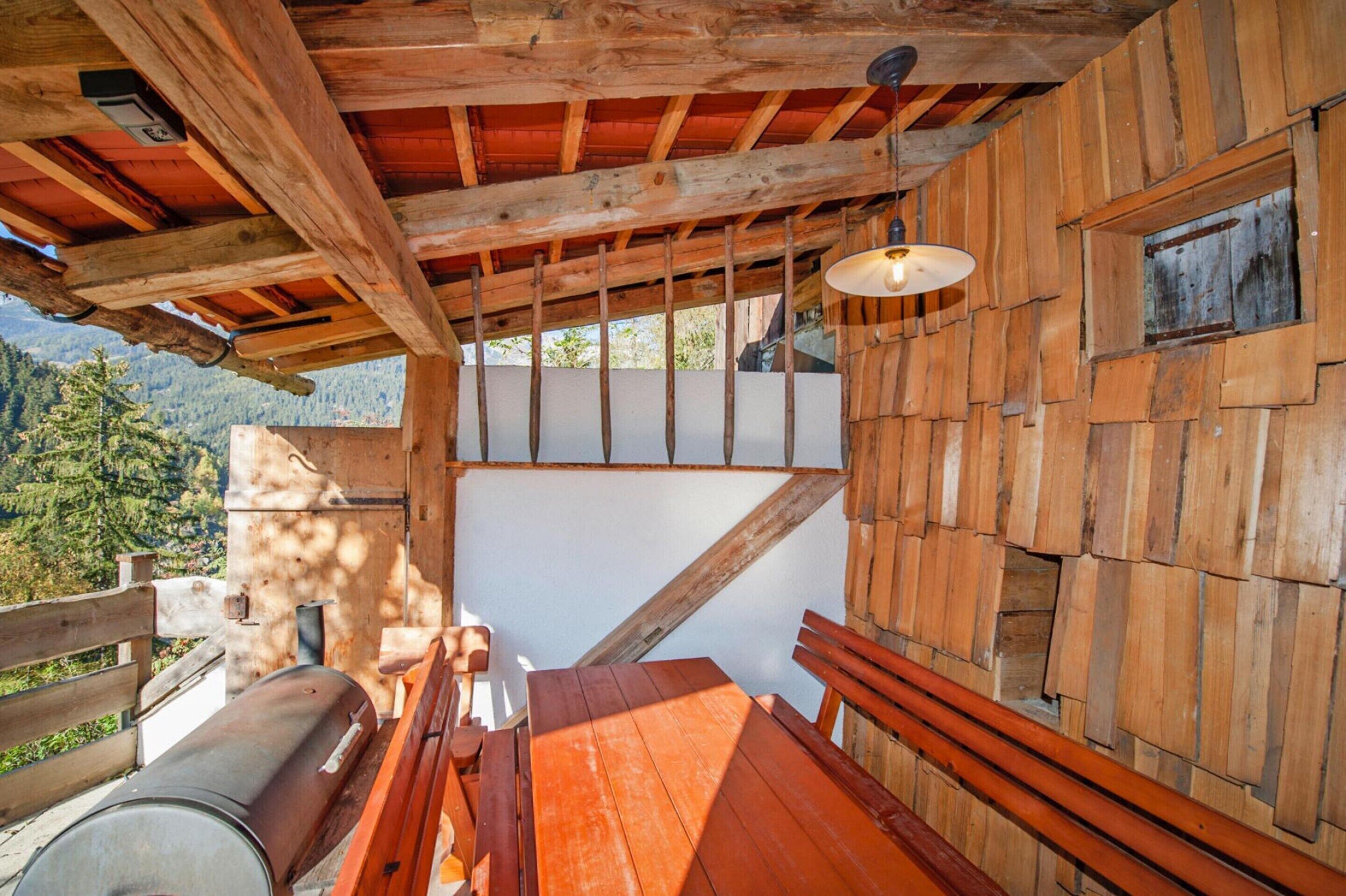 Une cabane en bois avec une table et des bancs en bois, un barbecue et une fenêtre donnant sur un paysage montagneux.
