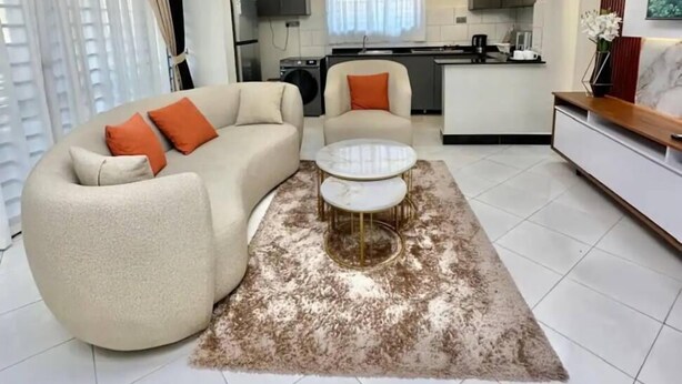 A modern living room with a beige sofa, two white round coffee tables, and a patterned rug. - LivLuxe Apartments 5 (Kampala)