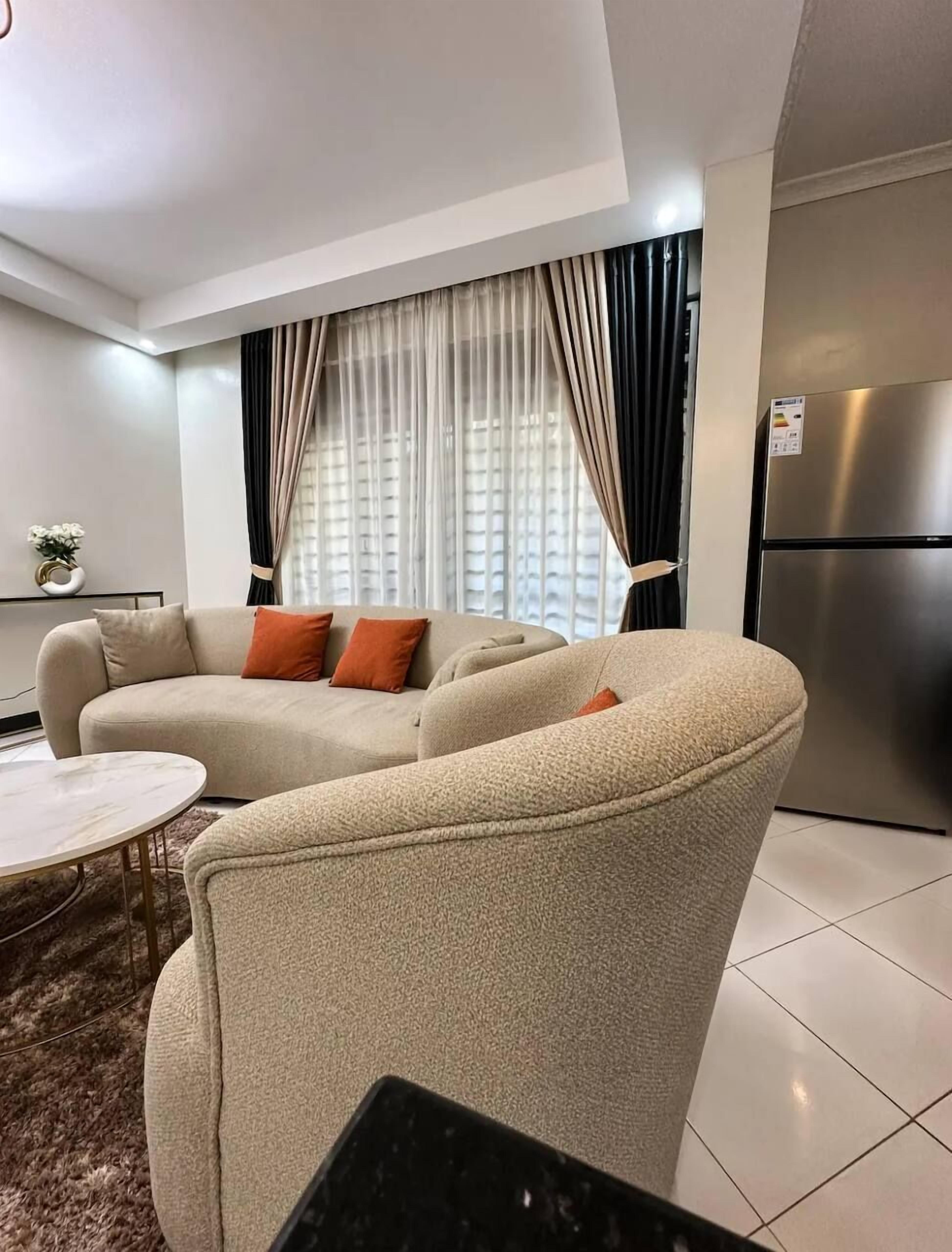 Un salon moderne avec un canapé d’angle beige, une petite table ronde et une grande fenêtre agrémentée de rideaux légers.